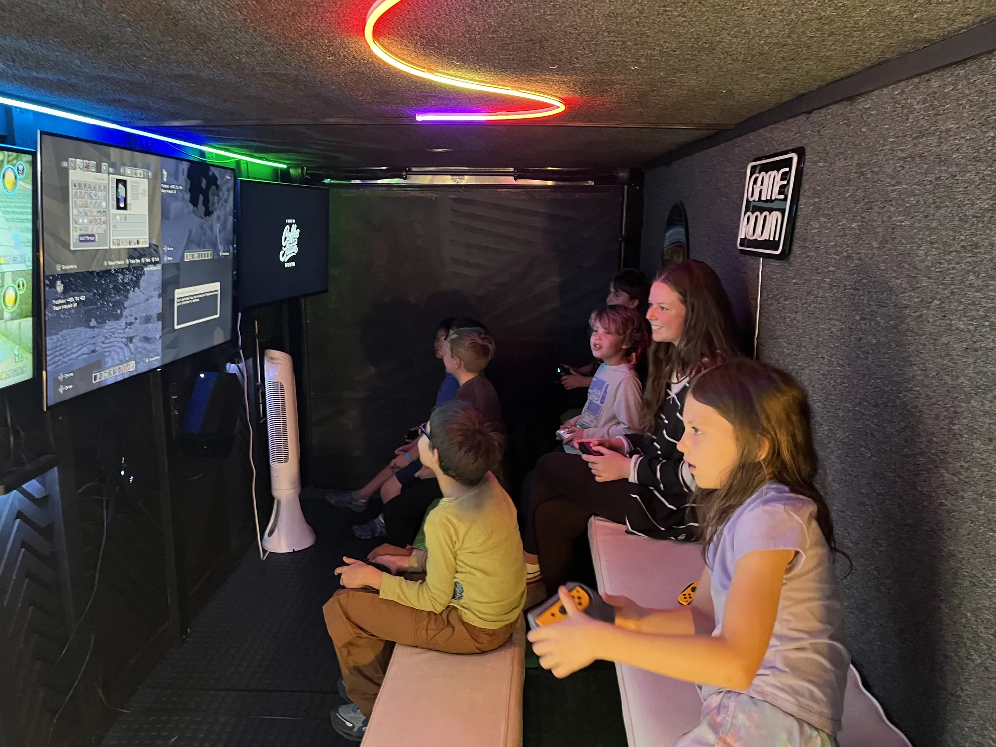 Birthday guests playing video games under the glowing Game Room sign inside the trailer