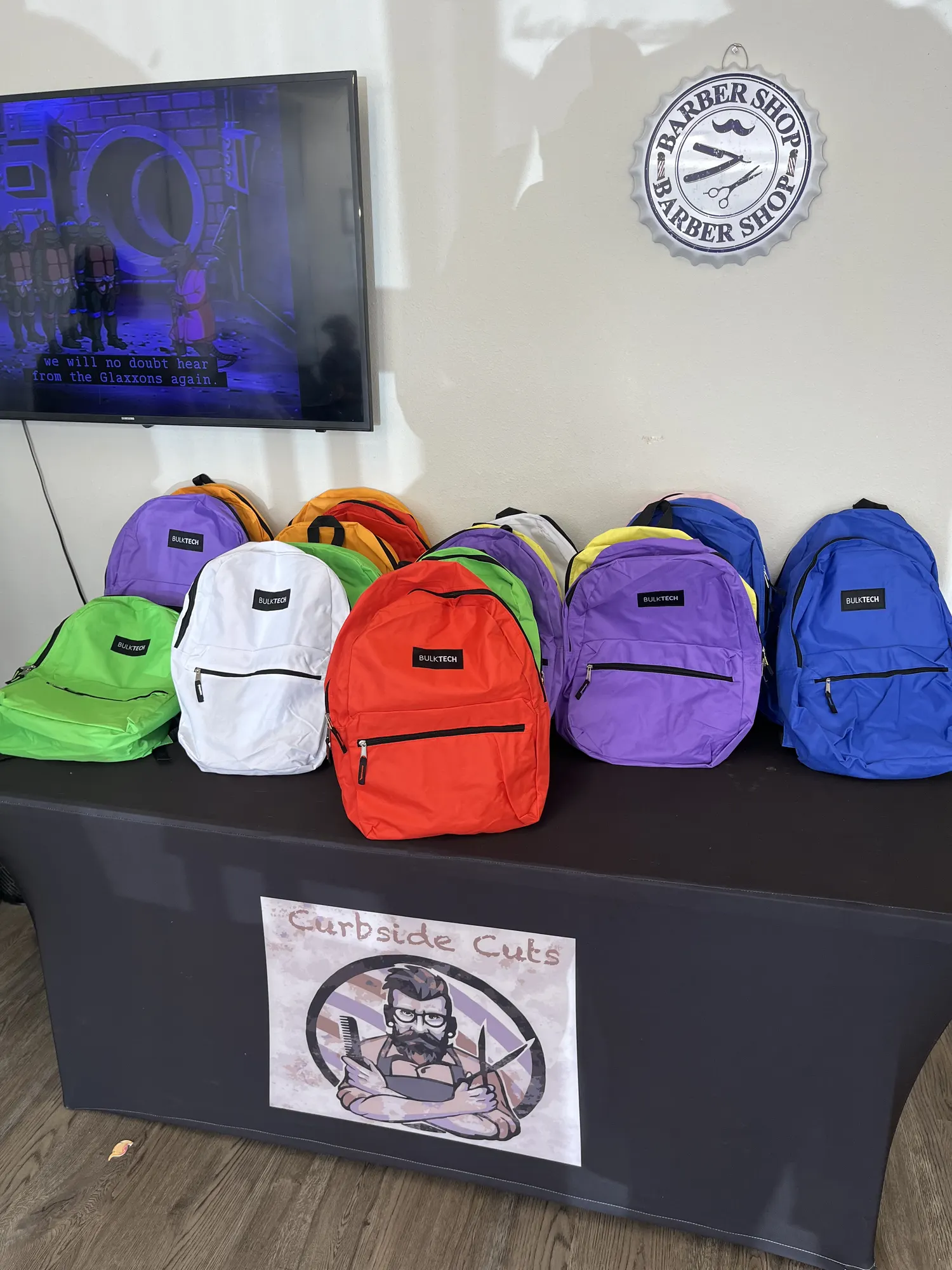 A table of bright new backpacks under a Curbside Cuts poster, ready for local kids