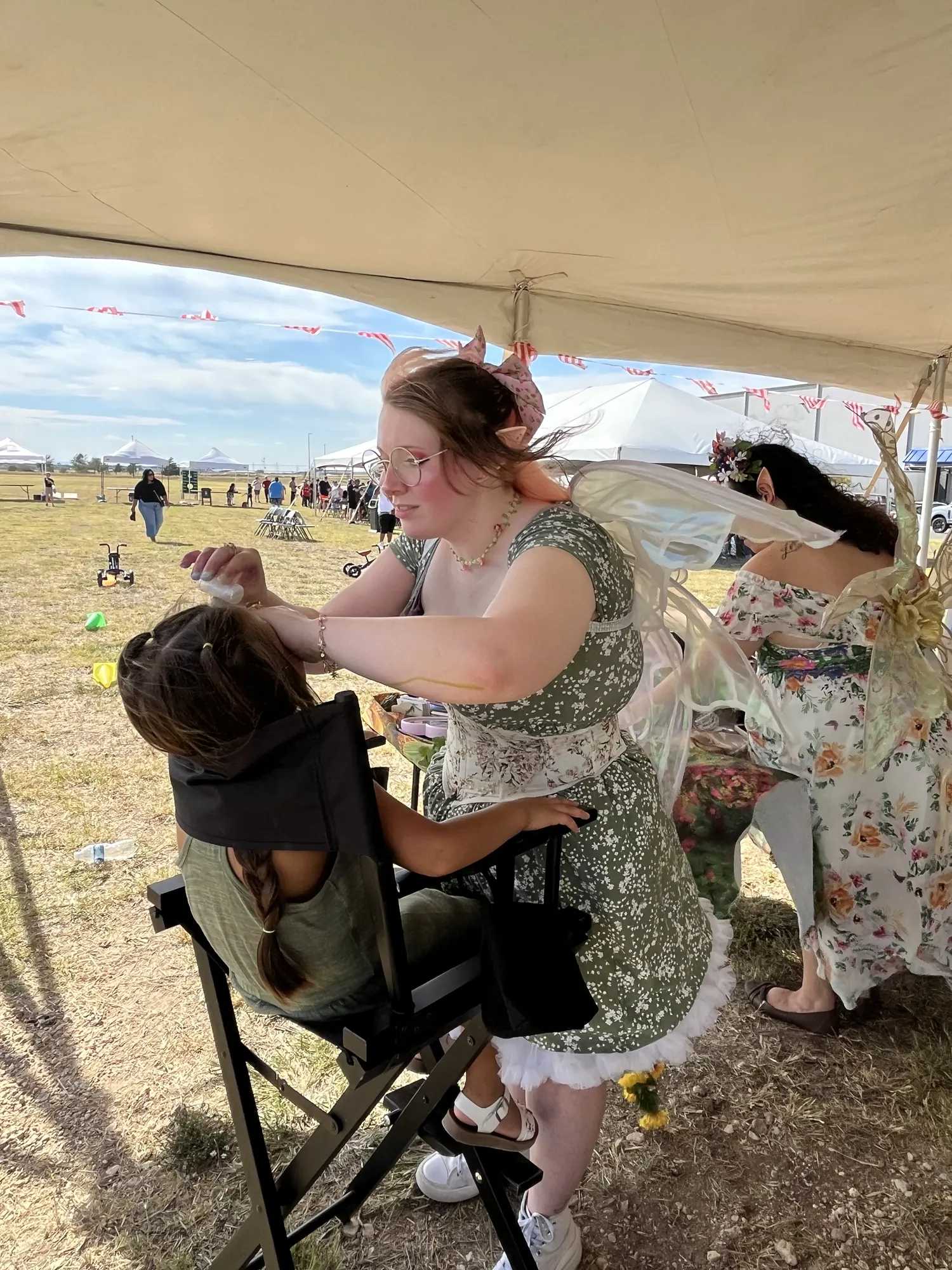 A face painter dressed as a fairy painting a child’s face under a tent at a company picnic in the Big Country