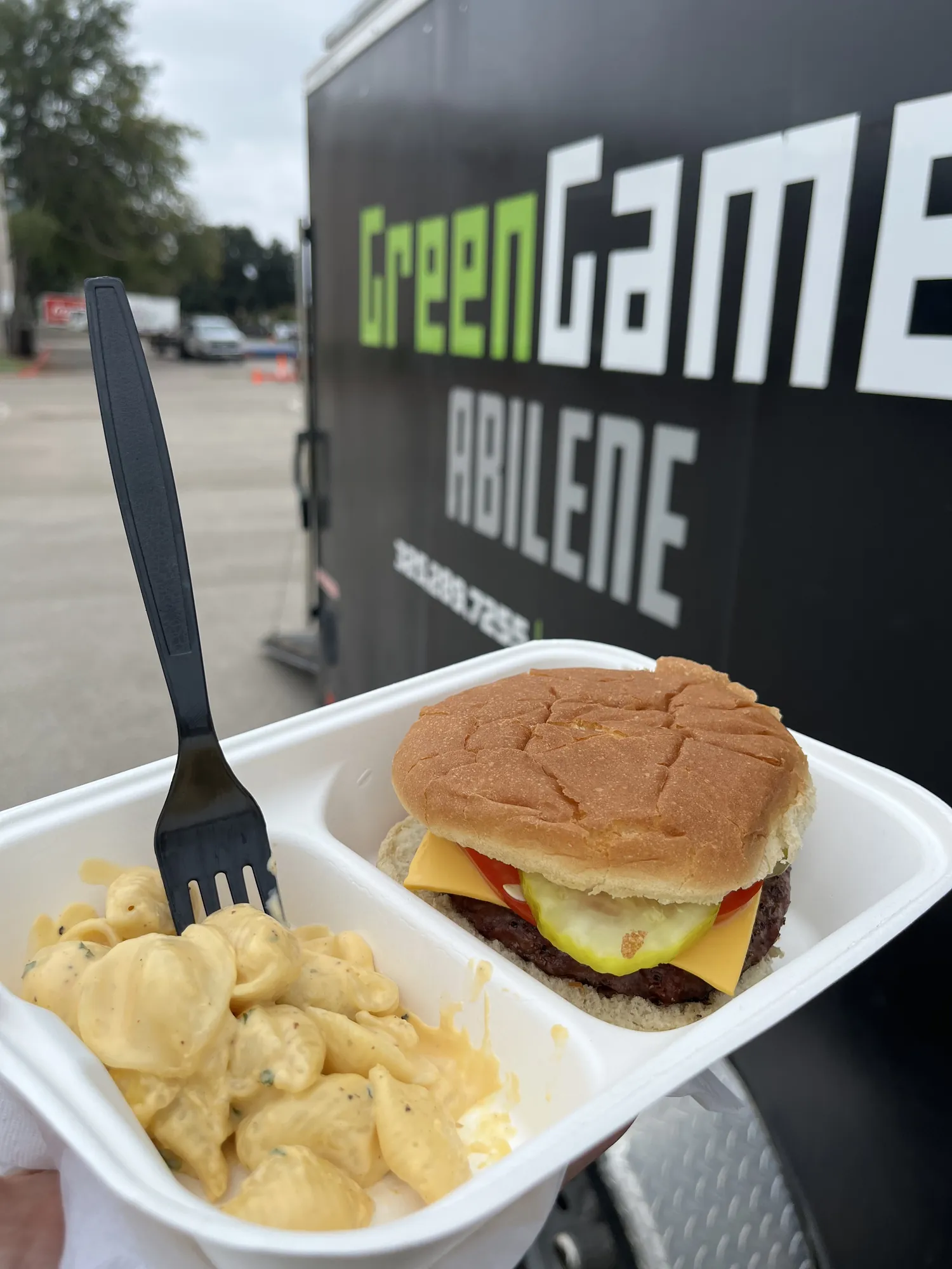 A cheeseburger and mac and cheese held up in front of the Green Games Abilene trailer