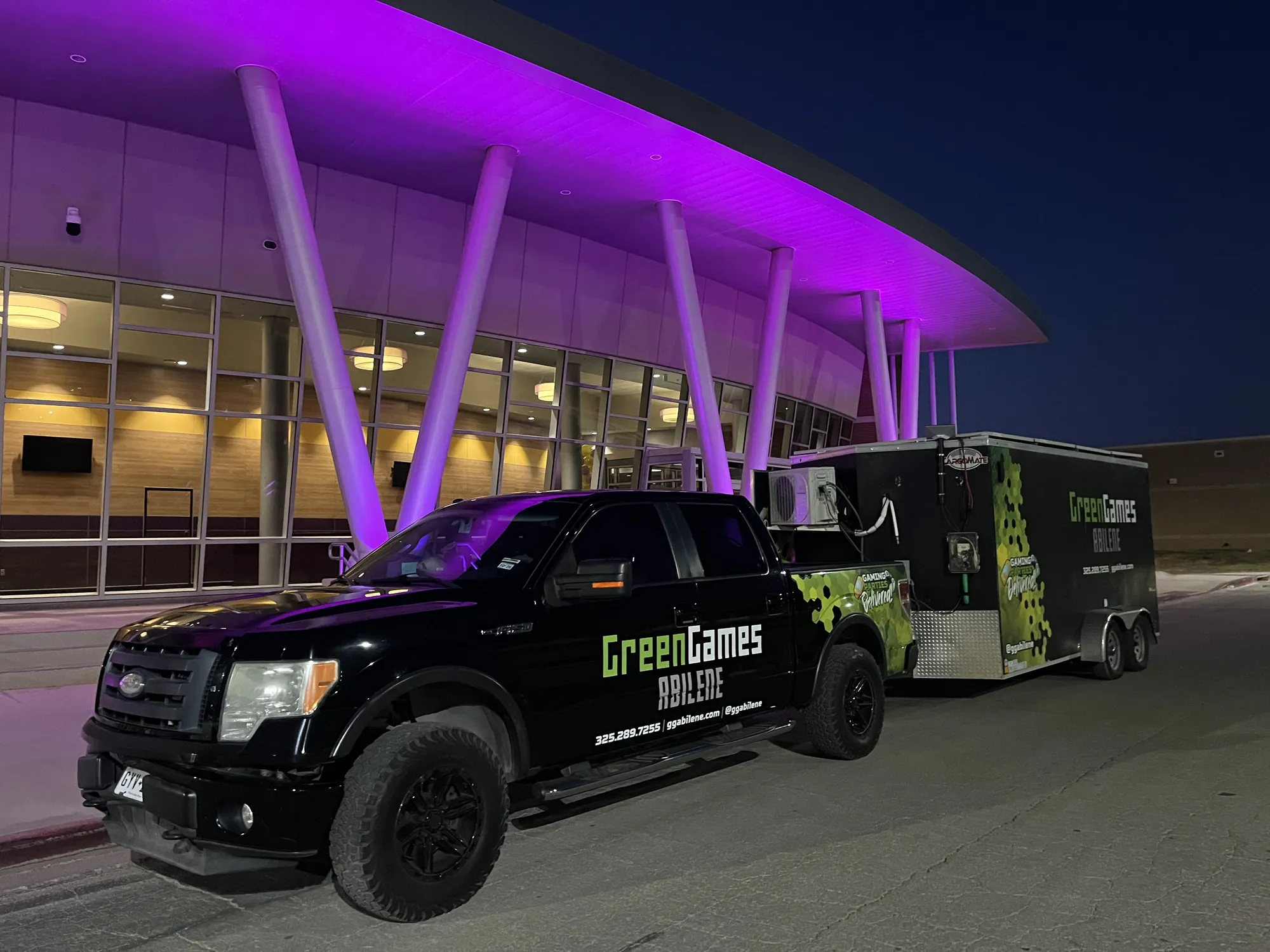 The Green Games Abilene truck and video game trailer parked outside a school lit up in purple at night