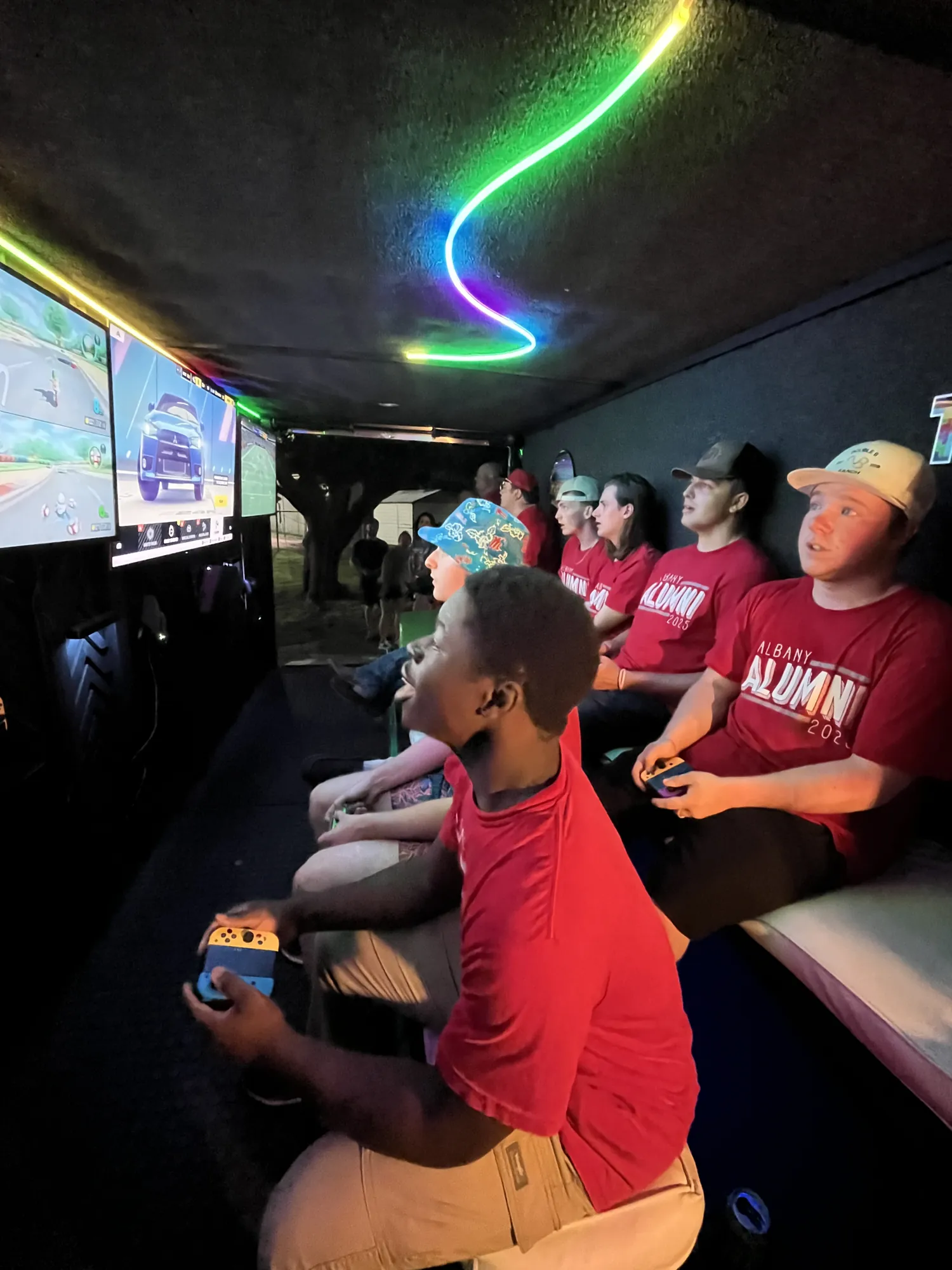 A graduate leaning into a racing game on the big screens under neon lights in the middle of the night